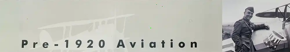 Two large exhibition display panels hung on a silver metal barrier system form a corner exhibition area. Text and graphics are shown on a neutral background. A large case contains a blue uniform jacket with matching hat and several small items including the first airborne transmitter, airplane models, instruments, and memorabilia.
