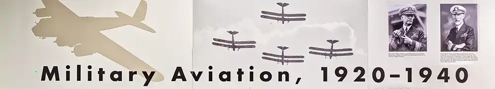 Two large exhibition display panels hung on a silver metal barrier system form a corner exhibition area. Text and graphics are shown on a neutral background. A large case contains airplane models, instruments, and uniform badges and insignia.