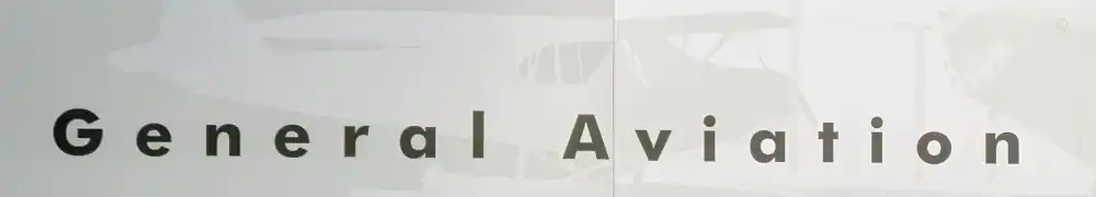 Two large exhibition display panels hung on a silver metal barrier system form a corner exhibition area. Text and graphics are shown on a neutral background. A large case contains two flight suits, airplane models, instruments, and memorabilia.