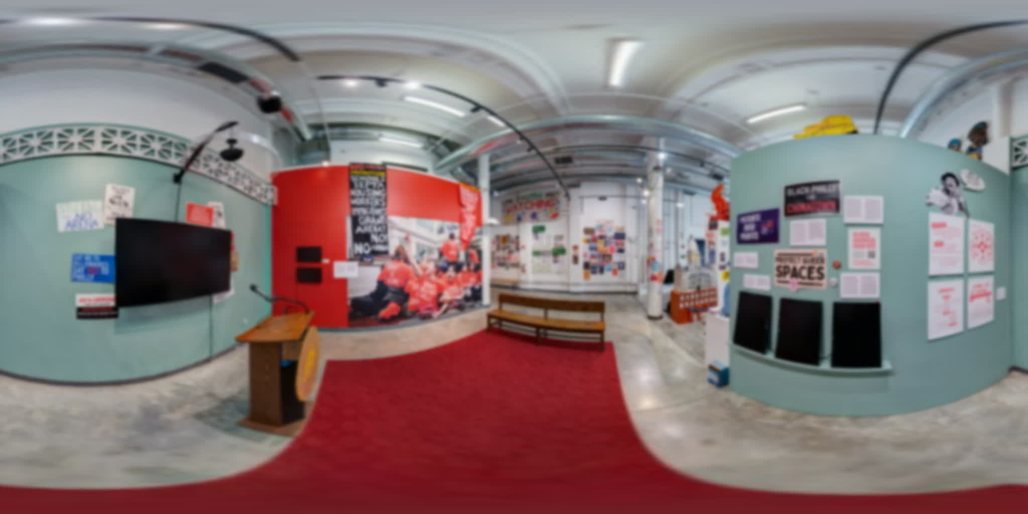 Teal and red gallery walls with a podium, six screens, protest signs, and a red carpet.