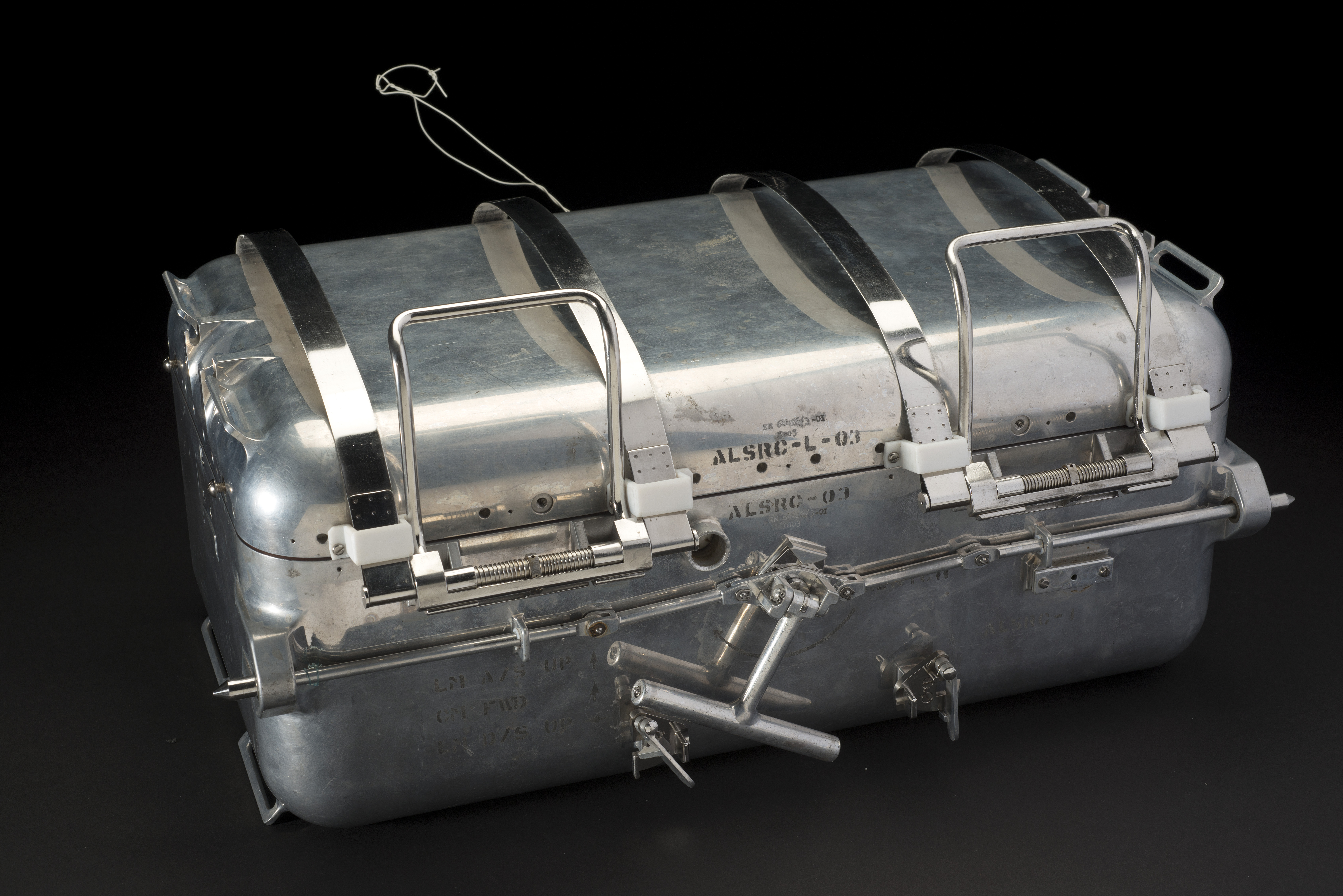 Bright aluminum sample case with four straps, two latches, a carrying bar, and identification lettering on the front.