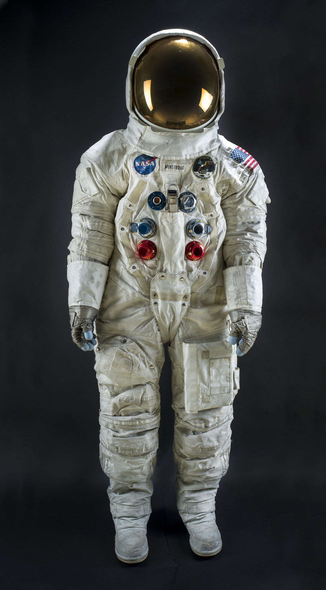 White multilayer Apollo lunar suit with helmet, gold visor, gloves, boots, chest connectors, pockets, flag, and mission patches.