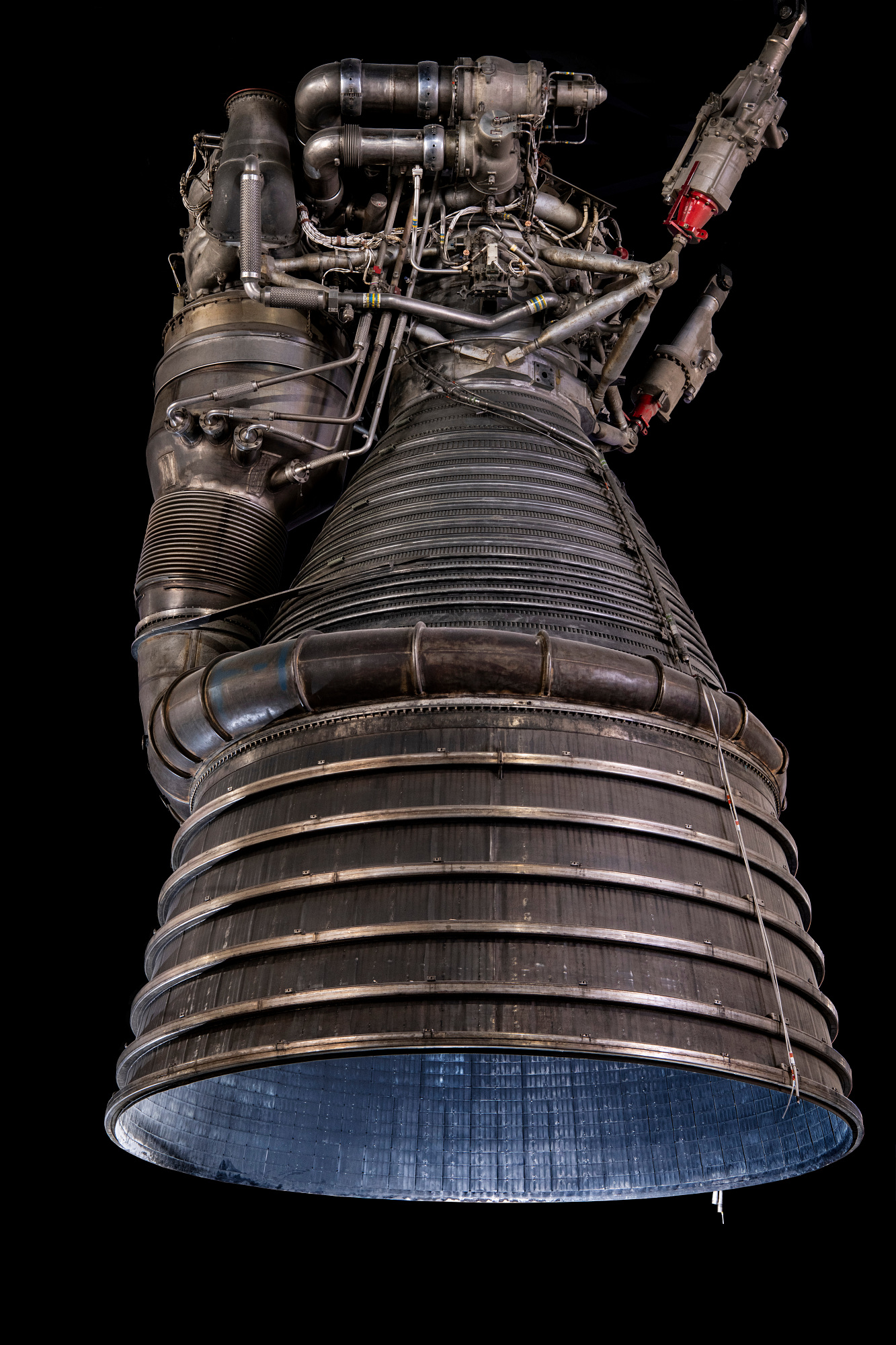 Large silver bell-shaped F-1 test engine with rings, pipes, structural supports, and a broad downward-facing nozzle.