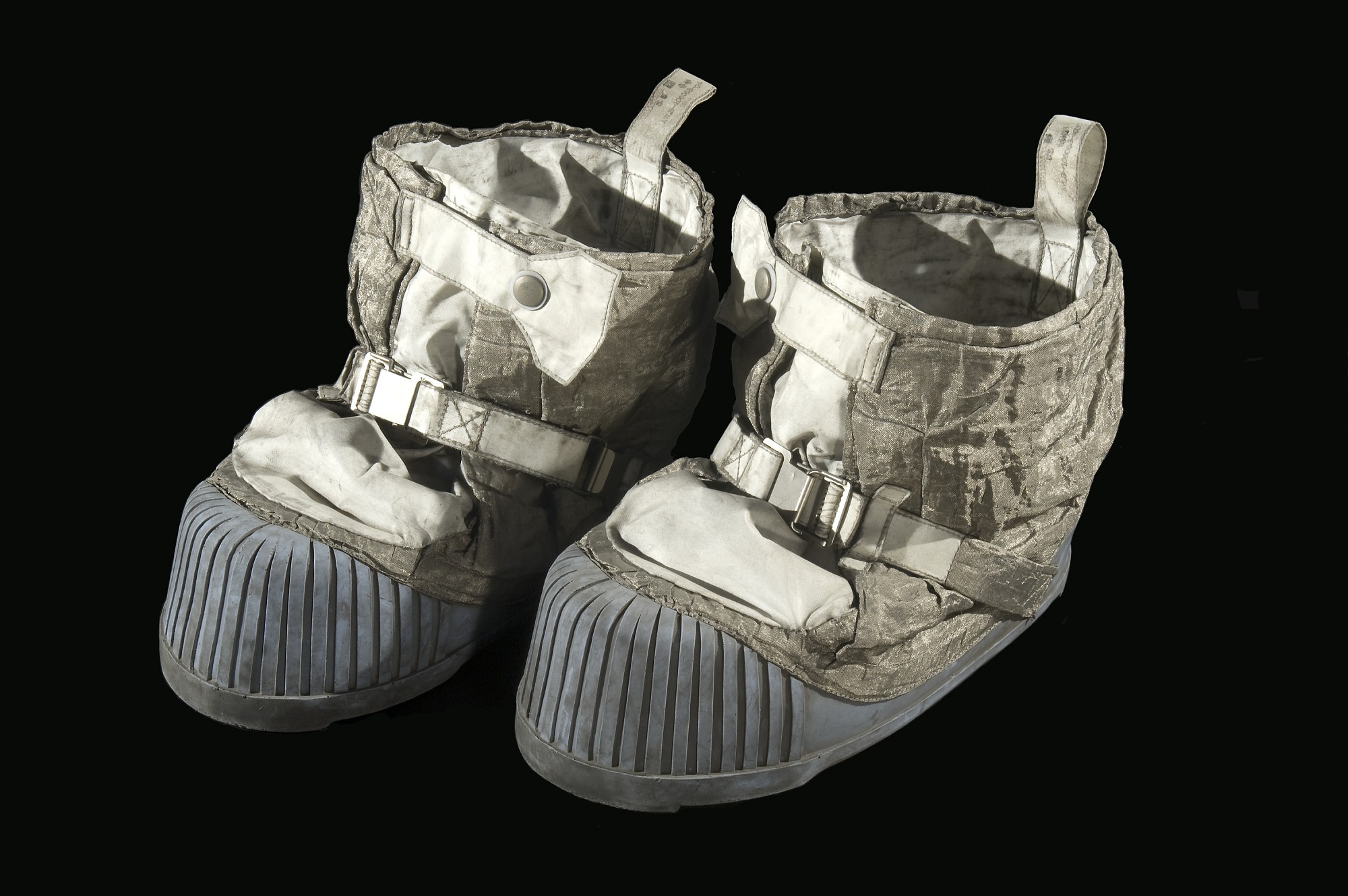 Pair of bulky white lunar overshoes with straps, buckles, loops, ribbed pale-blue soles, visible wear, and lunar dust.