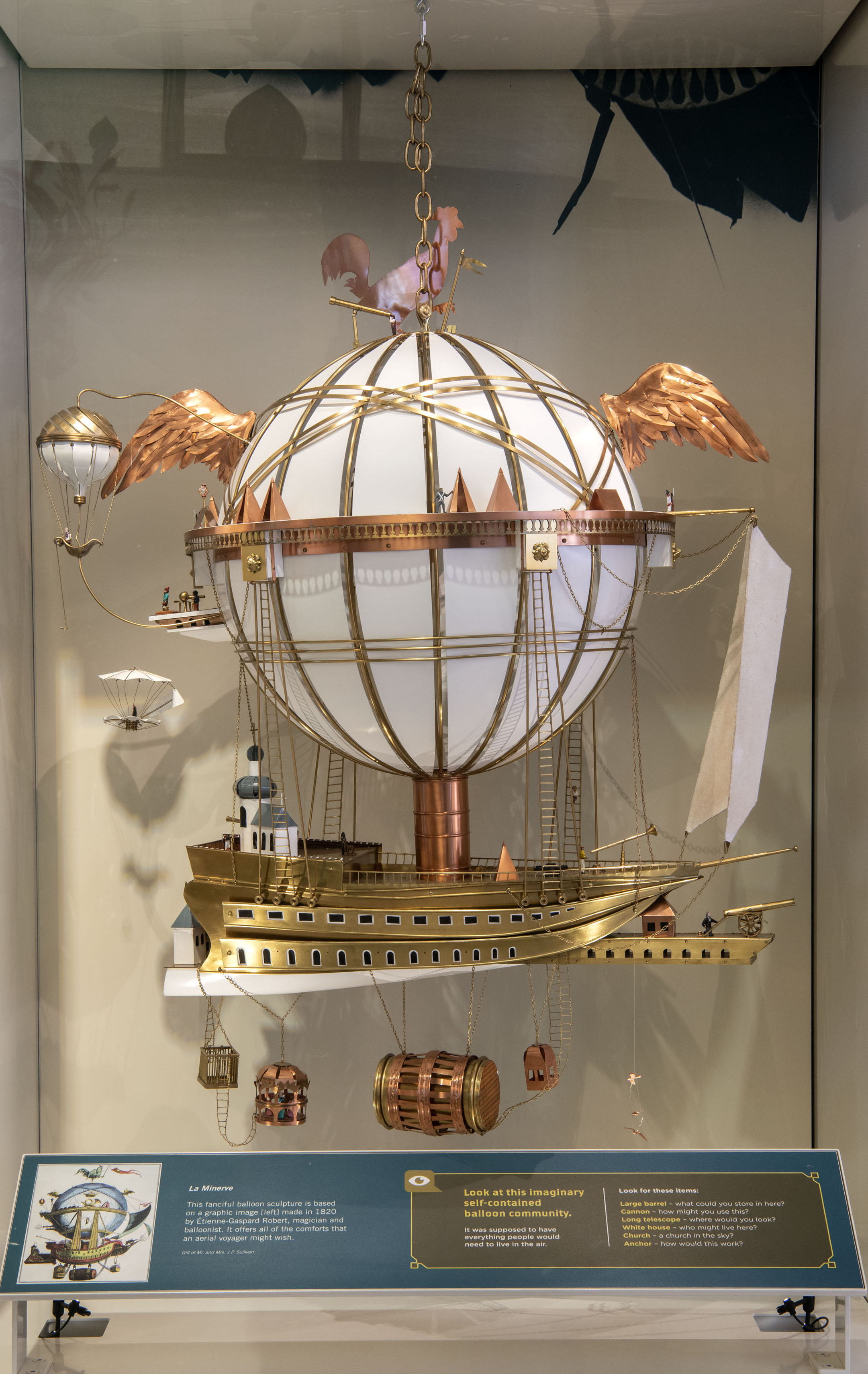 A fanciful balloon-and-ship sculpture with wings, observation decks, ladders, tents, and a copper rooster above the balloon.