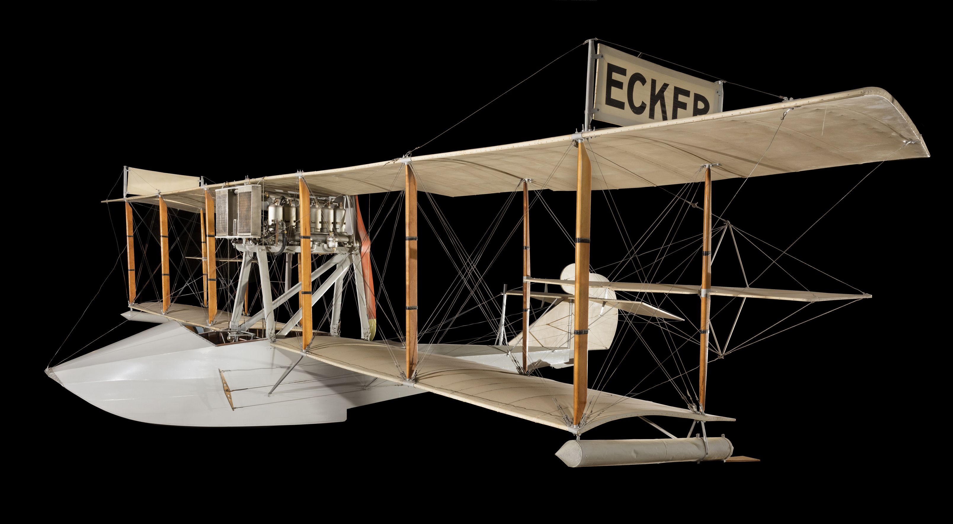 A light-gray wood-and-fabric biplane flying boat with a boat-shaped hull, engine between the wings, and ECKER marked above the upper wing.