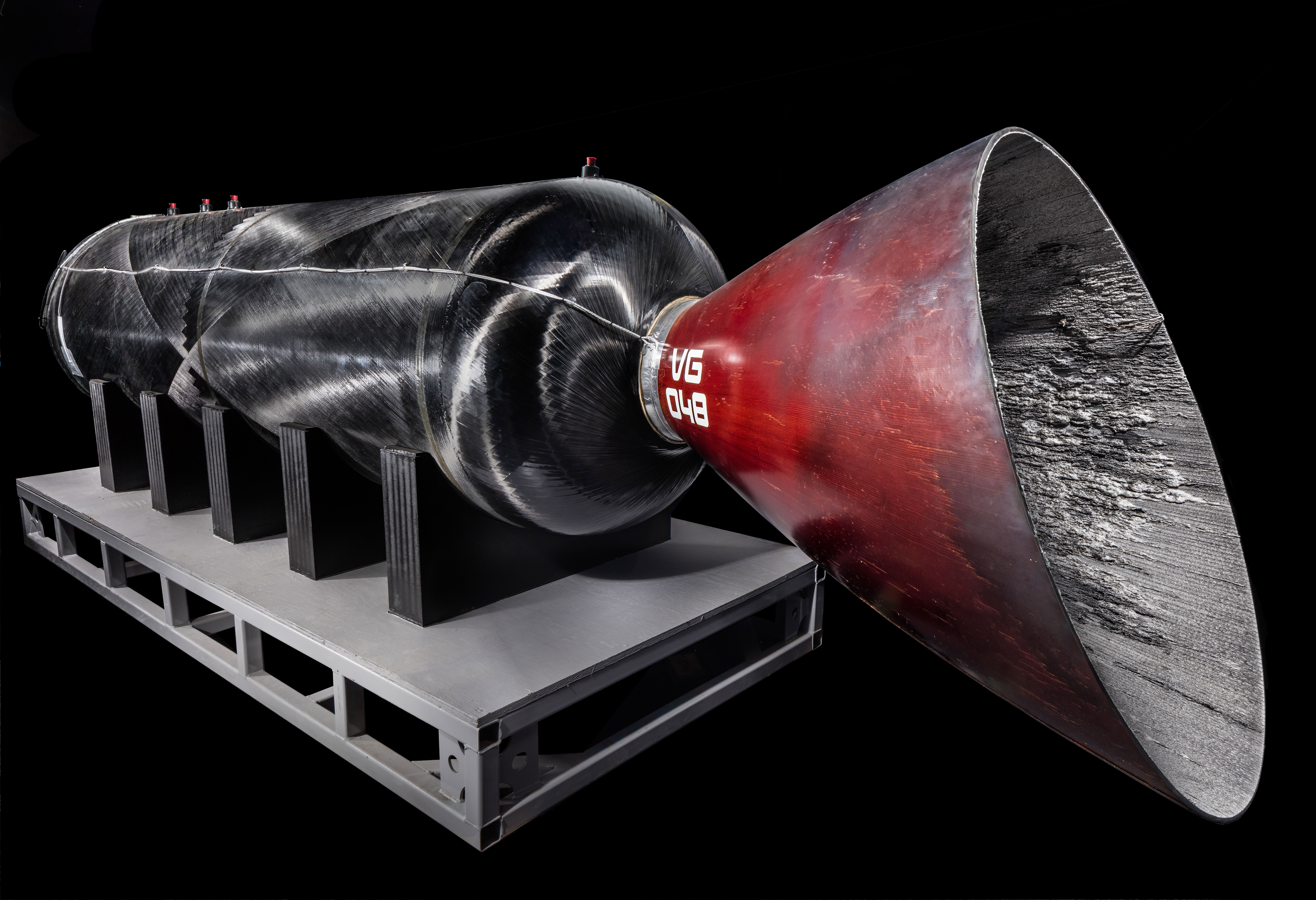 A long black cylindrical hybrid rocket engine displayed at an angle, with a red nozzle at one end.