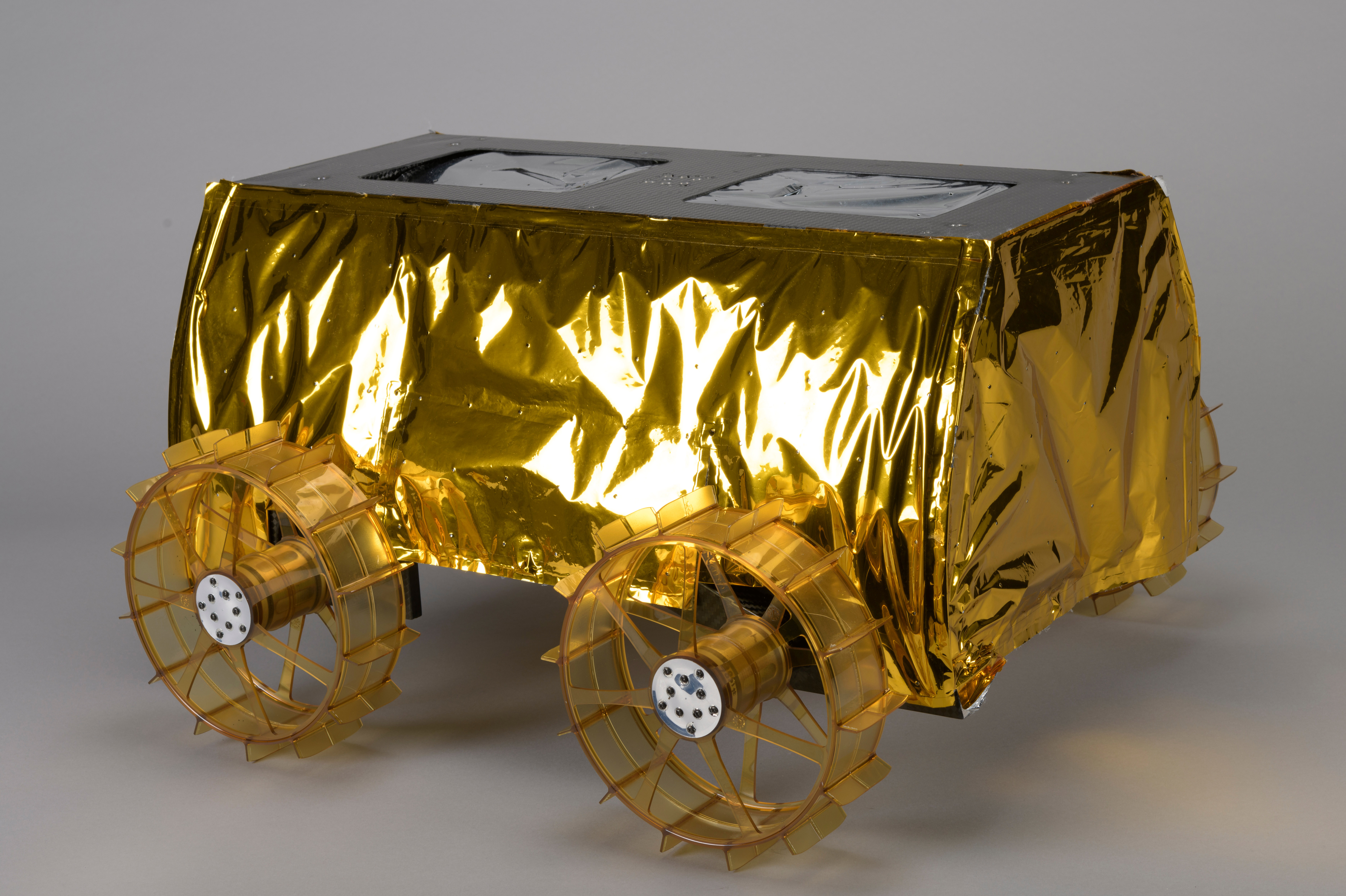 A compact box-shaped lunar rover wrapped in reflective gold foil, with four translucent spoked wheels and two rectangular panels on top.
