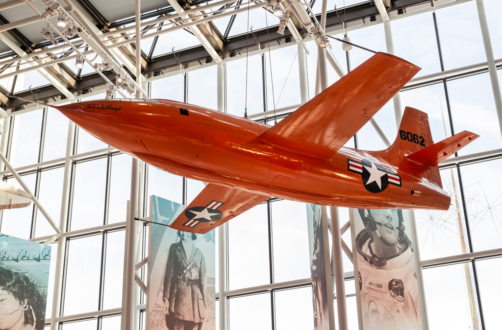 A small bright-orange experimental aircraft with a bullet-shaped fuselage, straight thin wings, and a clear cockpit canopy.