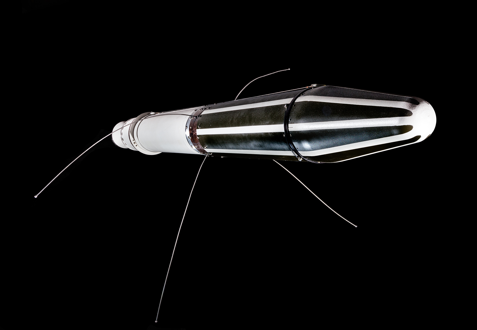 A long slender conical satellite with striped and white sections and four thin antennas, displayed near the spherical Sputnik 1.