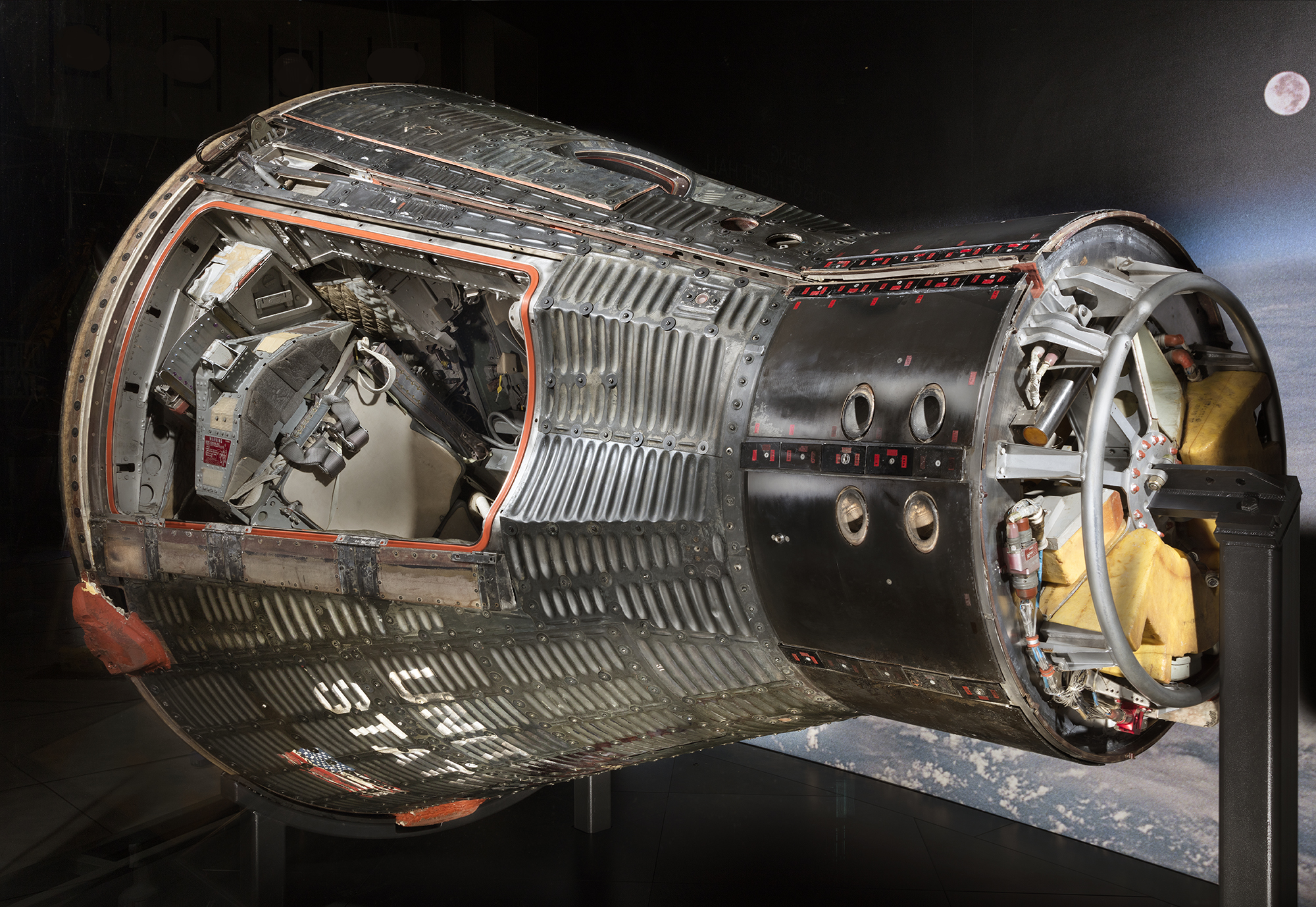 A corrugated-metal, cone-shaped two-person capsule displayed on its side with the hatch removed to reveal seats and controls.