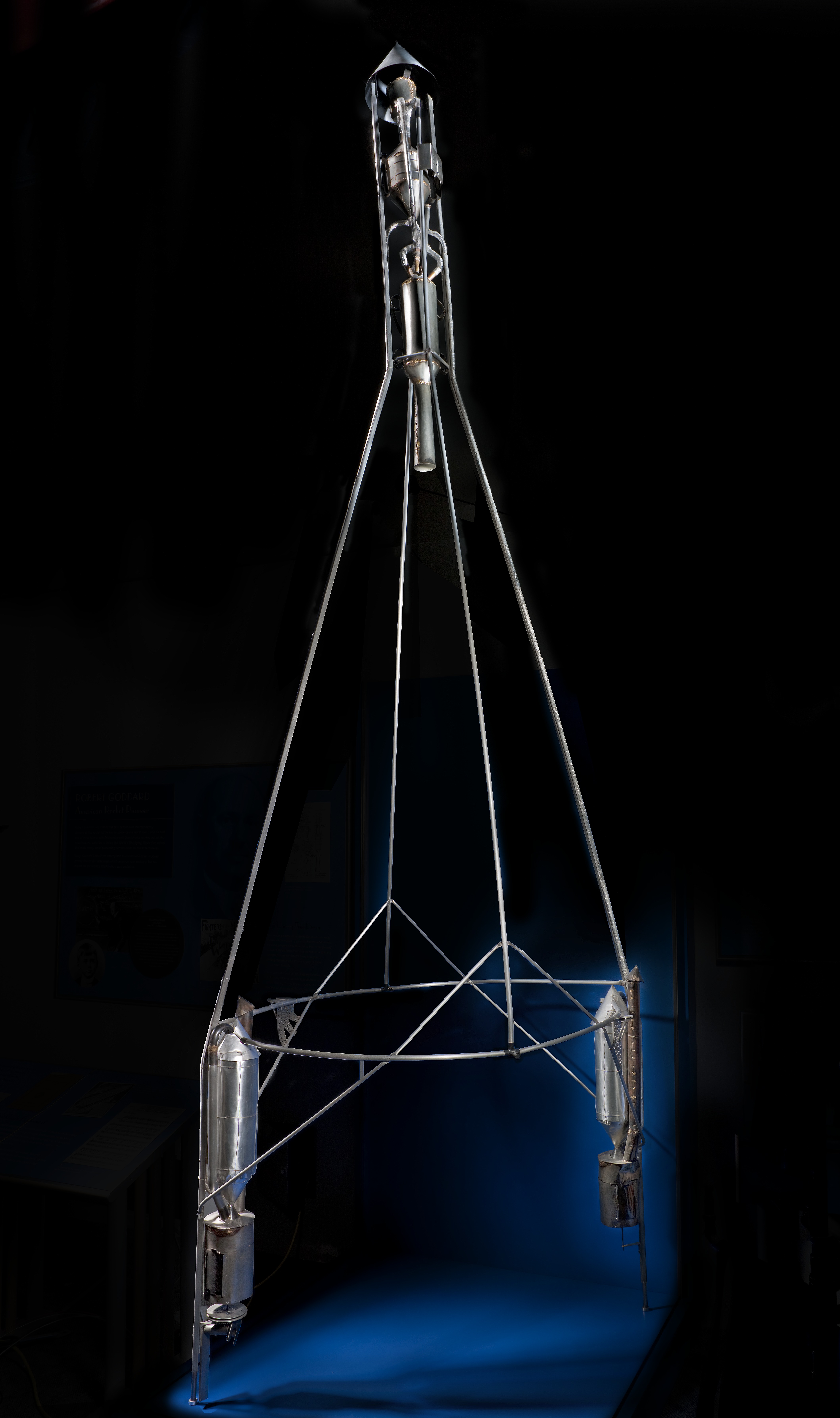 A tall open metal rocket framework with slender vertical poles, crossbars, cylindrical tanks, and a pointed top.