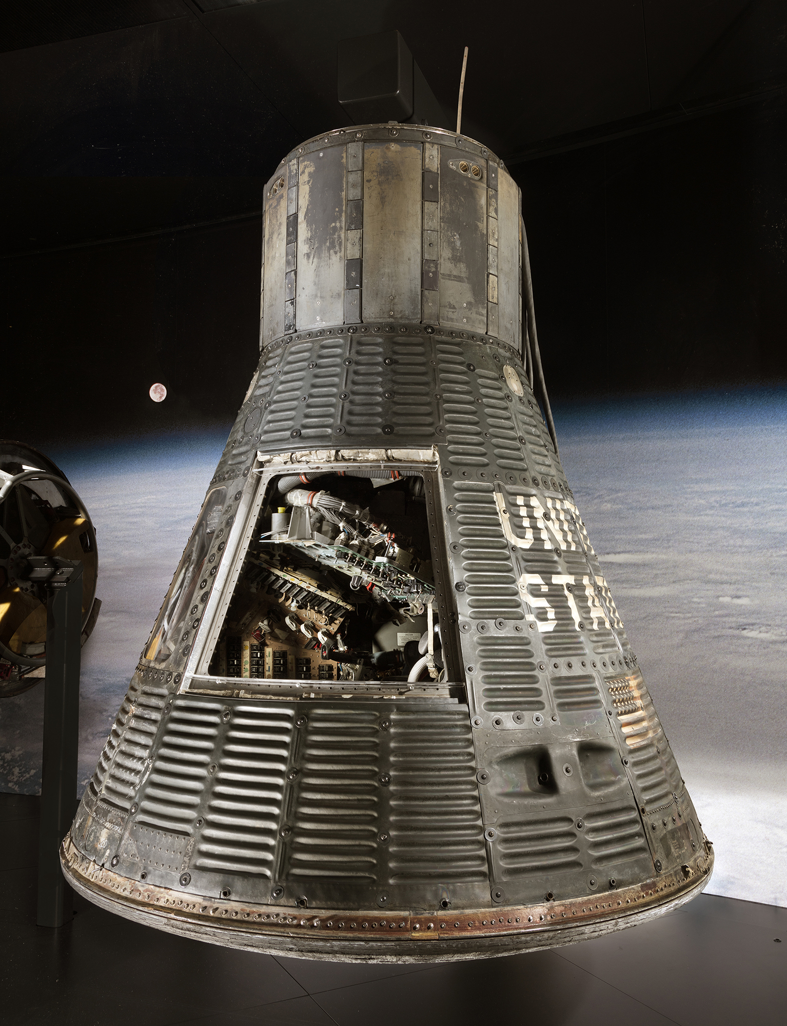 A corrugated-metal, cone-shaped one-person capsule displayed upright with its hatch removed to reveal a compact cockpit and instrument panel.
