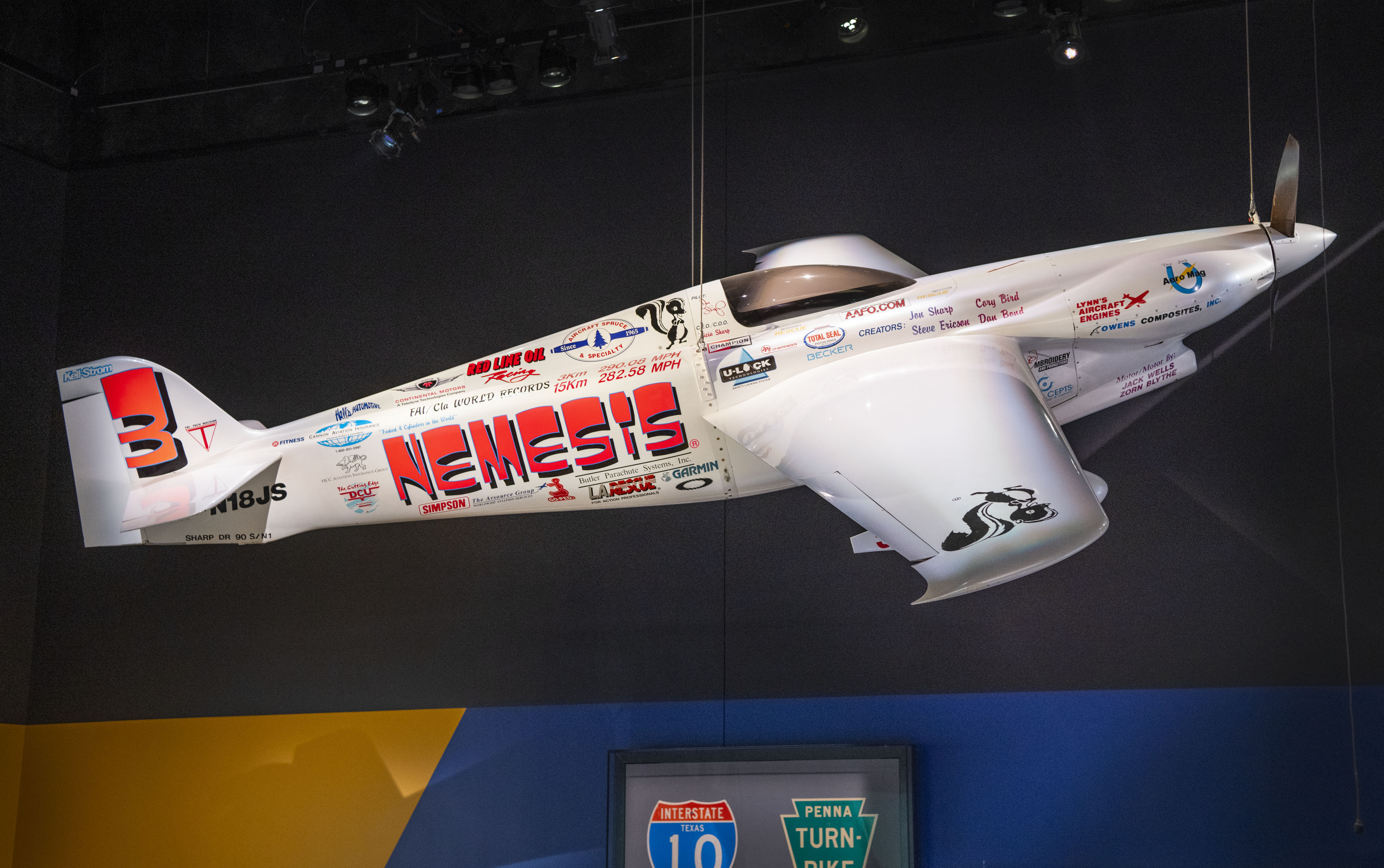 Small white single-seat racing aircraft with pink NEMESIS lettering, sponsor marks, and streamlined covered landing gear.