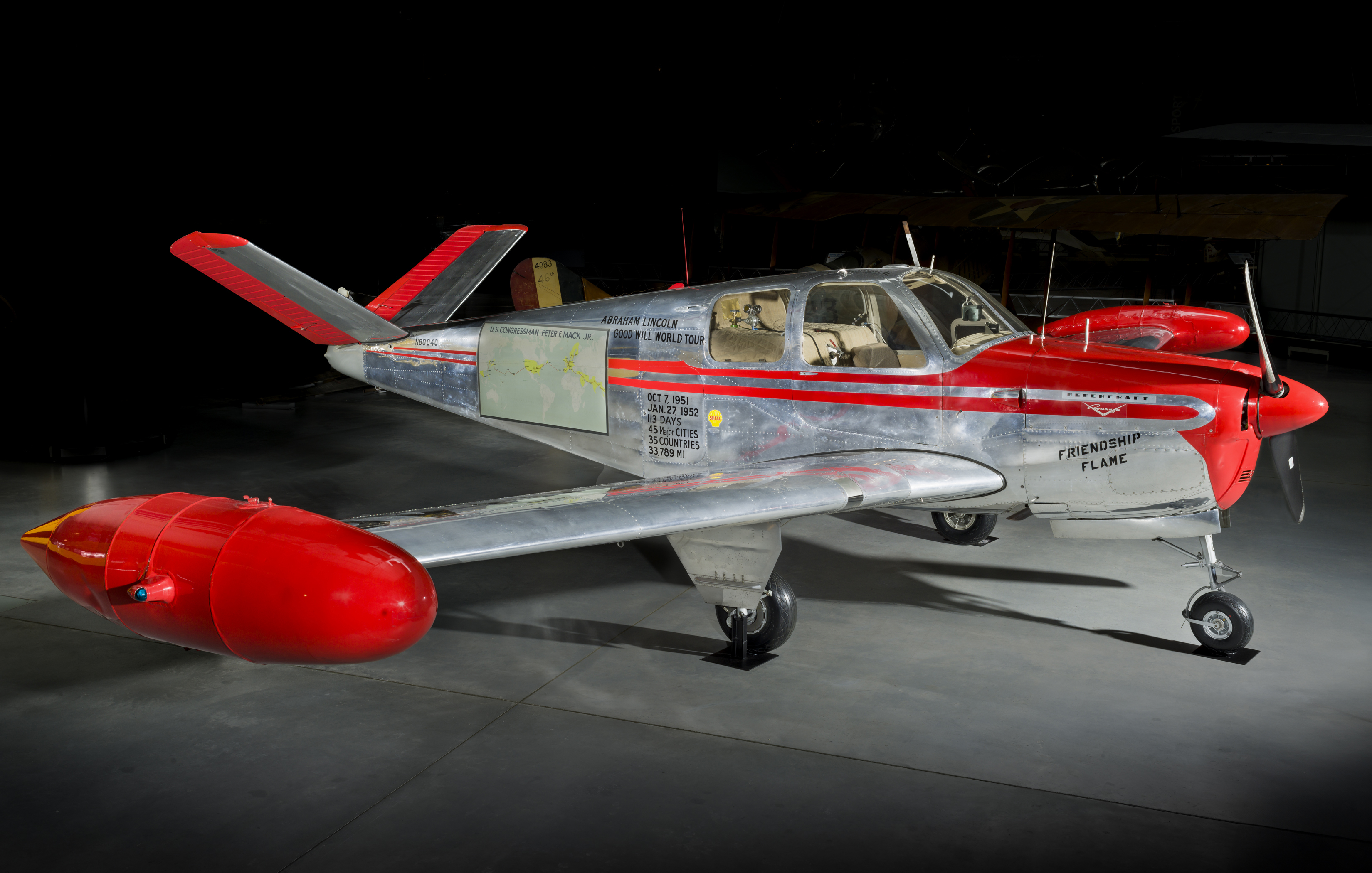 Silver and red four-seat airplane with a V-tail, tricycle landing gear, Friendship Flame markings, and a map of its goodwill tour.