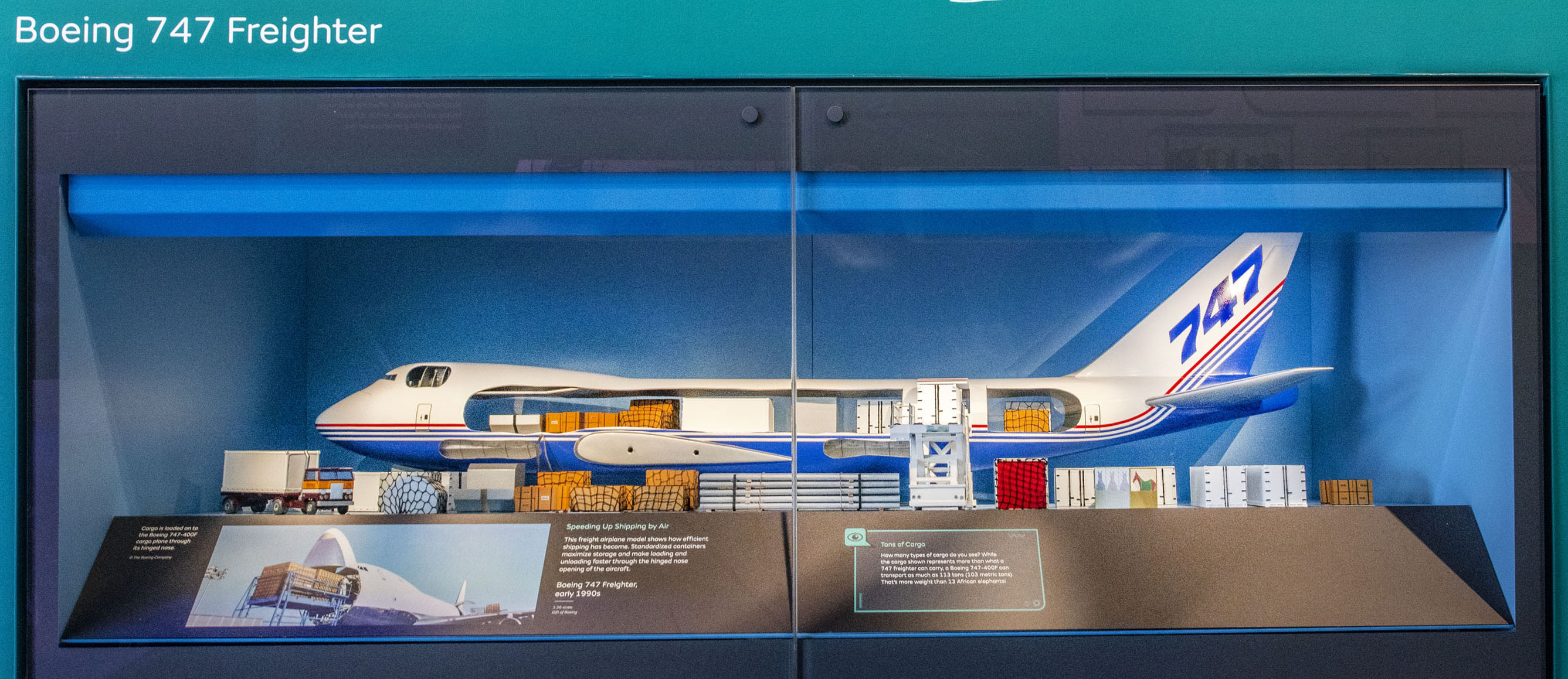 Cutaway model of a Boeing 747 freighter with its nose open, cargo containers arranged inside, and loading equipment beside it.