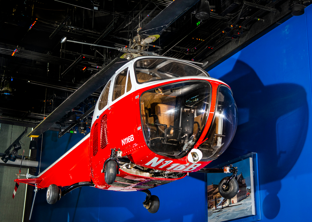 Red and white Bell Model 47B helicopter with a bubble cockpit, exposed structure, and wheeled landing gear.