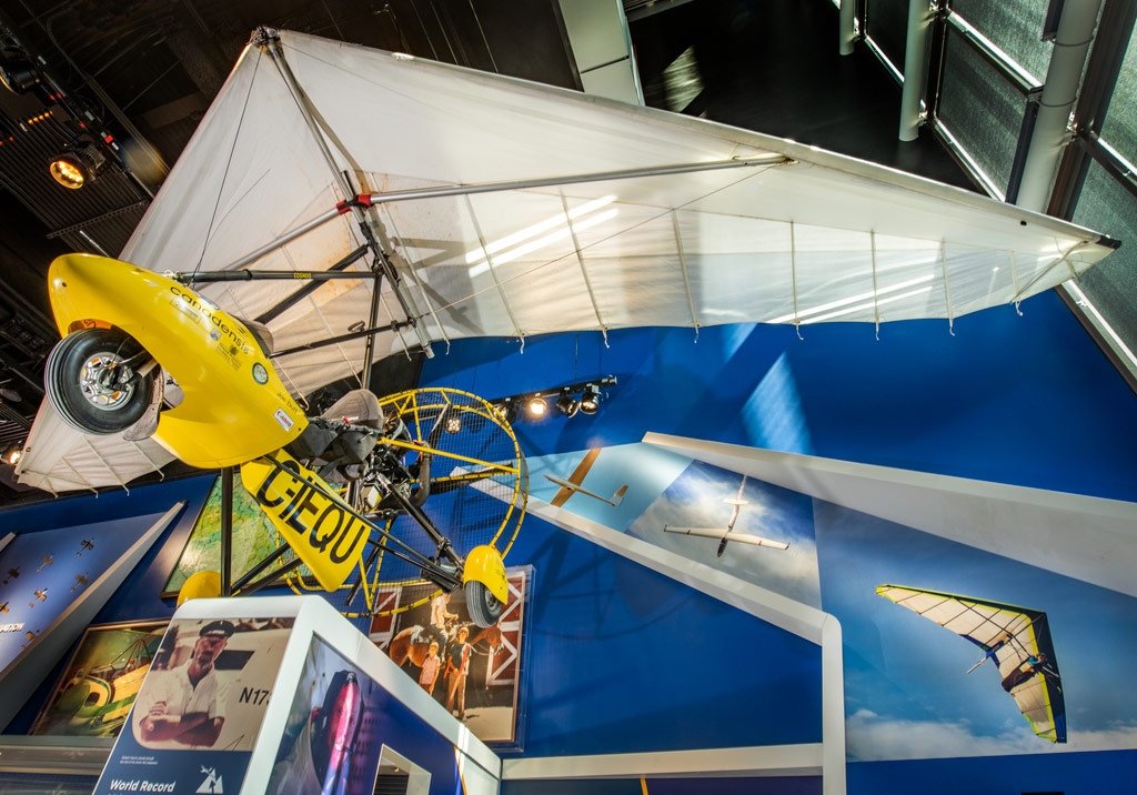 Yellow open-seat ultralight trike with a white Rogallo wing and a rear propeller inside a circular cage.