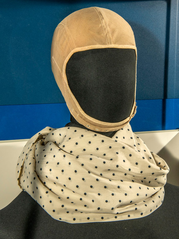 Beige cloth flight helmet and light silk scarf with dark polka dots displayed on a head form.
